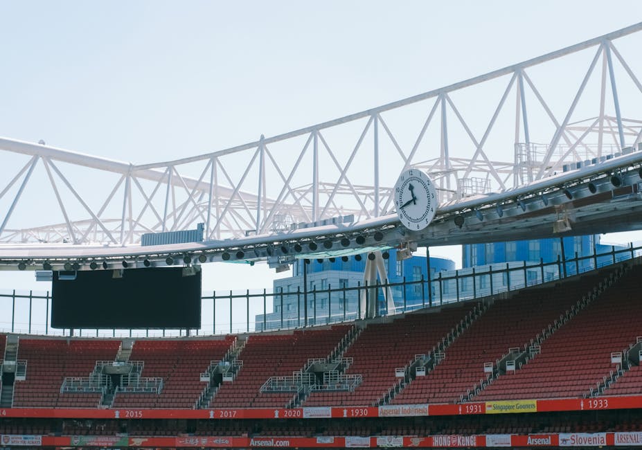 Inside a large sports stadium with red tiered seating, a section of the arena floor is visible, showing a row of empty chairs and clear pathways. In the background, a white steel structure with crisscrossing beams supports an overhanging roof. A round clock is mounted on the structure, displaying approximately 1:07. Near the seating area, several cardboard boxes, some wrapped in plastic or fabric covers, are being carefully loaded onto a moving trolley by a professional team. The boxes vary in size and are positioned close to the doorway leading outside. The interior environment is brightly lit, with natural light streaming through the stadium's open areas, highlighting the school or sports venue setting. This scene depicts a professional moving process related to event or venue relocation, with [COMPANY_NAME] handling the logistics of furniture and equipment transportation, typical of home or venue removals or packing and moving services, aligning with the theme of avoiding hidden charges during such operations.