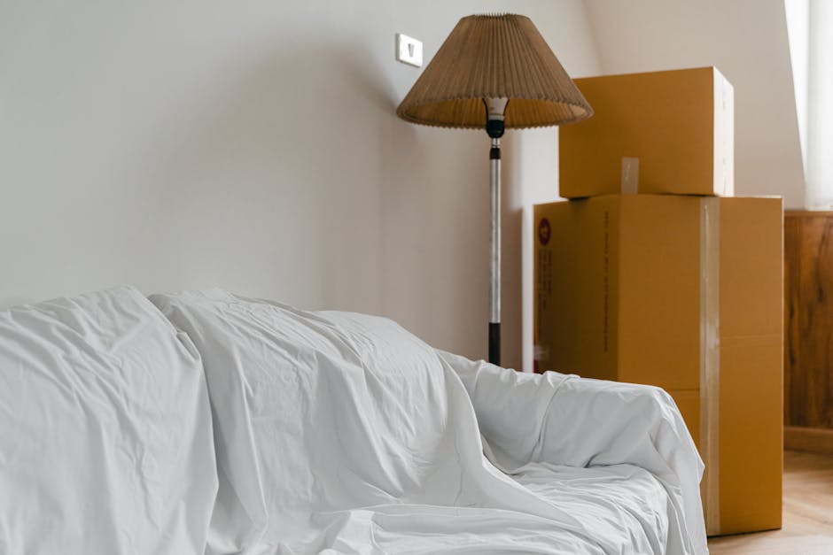 Inside a residential property, several packed cardboard boxes are stacked near the corner of a room, with three of varying sizes placed one on top of the other, ready for home relocation. To the left, a white fabric-covered sofa is visible, with some slight creases in the upholstery. A tall standing lamp with a brown pleated lampshade is positioned close to the boxes, casting soft light on the scene. The background features a plain wall with a small electrical switch and a window out of view, allowing natural light to illuminate the space. The arrangement suggests a packing and moving process, with the boxes prepared for transport by a professional home removal service such as Highbury Man and Van. The scene portrays the organized stage of packing furniture and household items ahead of a house move, highlighting typical elements involved in furniture transport and loading within a domestic environment.