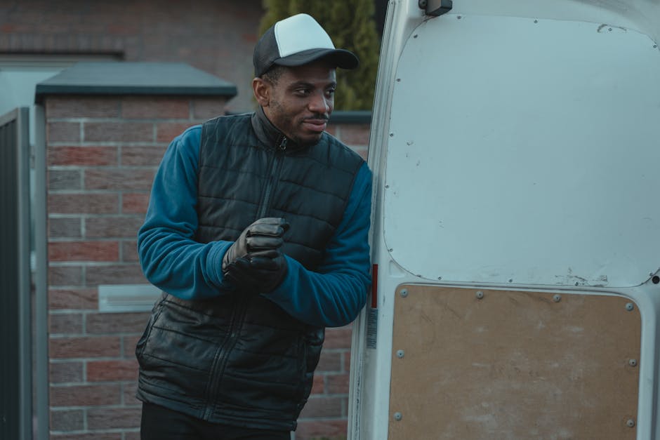A man wearing a baseball cap, dark gloves, a blue long-sleeve shirt, and a black padded vest stands outside a white van with an open rear door. He is positioned next to the loading area, which is on a paved driveway adjacent to a brick wall. Inside the van, visible packing materials include cardboard boxes and protective blankets, indicating a home relocation process. The man is holding a small object in his gloved hands while looking toward the van, possibly preparing for furniture transport or packing tasks. In the background, there are some trees and greenery, with natural lighting suggesting early evening or late afternoon. The scene captures the readiness and preparation involved in house removals, with [COMPANY_NAME] providing professional moving services related to packing, loading, and furniture transport as part of a complete house removal operation.