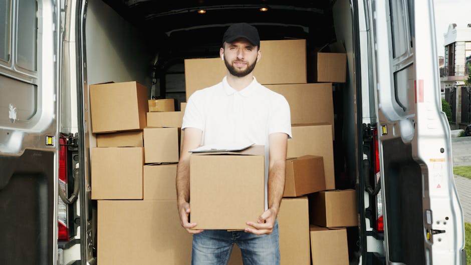 A professional mover from Highbury Man and Van stands at the rear of a large, open van filled with stacked cardboard boxes of various sizes, some wrapped in plastic or packing paper, ready for home relocation. The van is parked outside a residential area with houses and greenery visible in the background, and the image is taken during daylight with natural lighting. The mover, dressed in casual attire including a white polo shirt, jeans, and a black cap, holds a medium-sized box in front of him, with a clipboard resting on top of it. The inside of the van shows a spacious cargo area with additional boxes securely arranged to facilitate furniture transport and packing and moving processes, emphasizing the careful loading typical during a house removal. The scene captures the logistical steps involved in household furniture transport, with focus on the materials, spatial arrangement, and equipment used by [COMPANY_NAME] in relocation services.