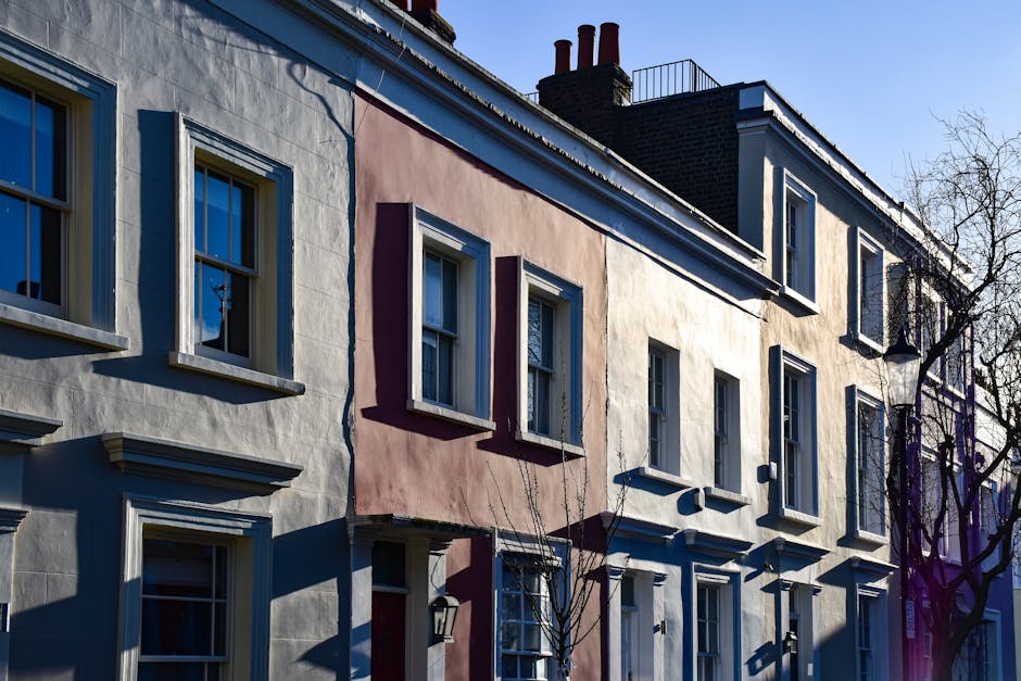 A row of terraced houses with light-colored facades in a residential area, showing multiple windows with white frames. The houses are located on a street with a clear blue sky above, and the sunlight casts shadows on the building facades. Visible on the right side is a leafless tree with branches extending toward the sky. In front of the houses, a delivery van is parked on the pavement, partially visible on the right, with its rear doors open for loading or unloading. Several cardboard boxes, wrapped with plastic film, are arranged on the pavement beside the van, suggesting a home relocation or furniture transport process. An individual, wearing work gloves and casual clothing, is engaged in lifting or carrying a box toward the van, involving a typical furniture transport or packing and moving activity. The scene captures a moment of the loading process, with additional packing materials and blankets possibly present for protecting furniture during the move, and is indicative of professional removals services supported by Highbury Man and Van, in the context of house removals in Highbury.