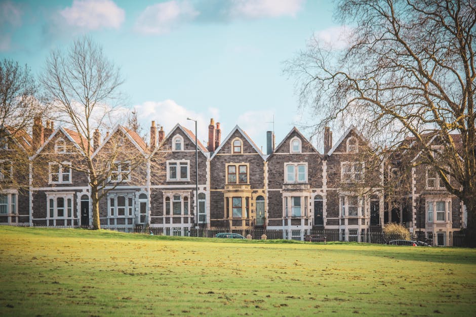 A row of Victorian-style terraced houses with grey brick facades and white trim, set behind a grassy park area with leafless trees, under a partly cloudy blue sky. In the foreground, there is a paved pathway leading up to the properties. No furniture or moving equipment is visible in the image. This scene depicts the exterior environment of a typical residential area, suitable for house removals or relocation services, such as those provided by Highbury Man and Van, while focusing on the external view rather than inside the properties.