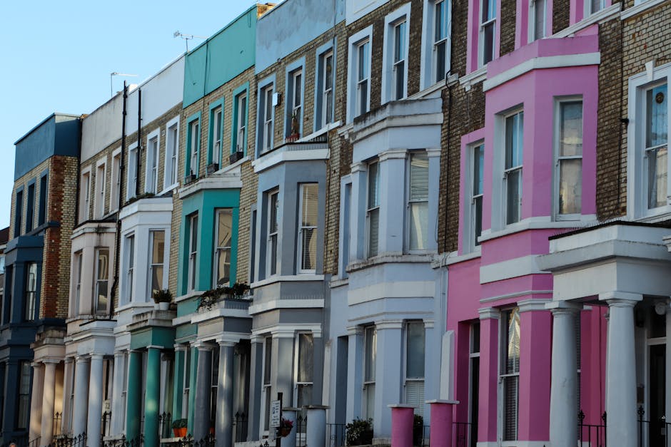 A row of terraced houses with light-colored facades in a residential area, showing multiple windows with white frames. The houses are located on a street with a clear blue sky above, and the sunlight casts shadows on the building facades. Visible on the right side is a leafless tree with branches extending toward the sky. In front of the houses, a delivery van is parked on the pavement, partially visible on the right, with its rear doors open for loading or unloading. Several cardboard boxes, wrapped with plastic film, are arranged on the pavement beside the van, suggesting a home relocation or furniture transport process. An individual, wearing work gloves and casual clothing, is engaged in lifting or carrying a box toward the van, involving a typical furniture transport or packing and moving activity. The scene captures a moment of the loading process, with additional packing materials and blankets possibly present for protecting furniture during the move, and is indicative of professional removals services supported by Highbury Man and Van, in the context of house removals in Highbury.