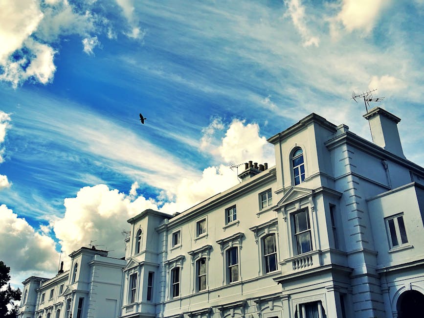 A white, multi-story residential building with classic architectural details, including decorative window surrounds and cornices, seen from below against a partly cloudy sky with blue and white hues. An individual or team from Highbury Man and Van is engaged in home relocation activities, loading or unloading a van parked near the building, carrying or arranging cardboard boxes, wrapped furniture, and packing materials such as plastic film and blankets. The scene captures the process of furniture transport and packing and moving preparations, with the van positioned close to an open doorway, in a residential street environment with clear lighting. The image illustrates house removals and the logistical aspects involved in house relocations, aligning with the topic of Islington Council permits for Highbury removals.