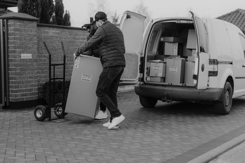 Two movers are loading a large cardboard box onto a hand truck outside a residential property, with a white cargo van parked nearby. The van's rear doors are open, revealing several packed cardboard boxes inside, secured with plastic wrap and packing tape. One mover is wearing a baseball cap and gloves, guiding the box onto the hand truck, while the other assistant helps lift and position the item. The scene takes place on a paved driveway adjacent to a brick wall and a garden area, under overcast skies, during daylight hours. This image illustrates a home relocation process involving furniture transport, packing, and moving logistics handled by Highbury Man and Van, with attention to careful handling and proper loading procedures.