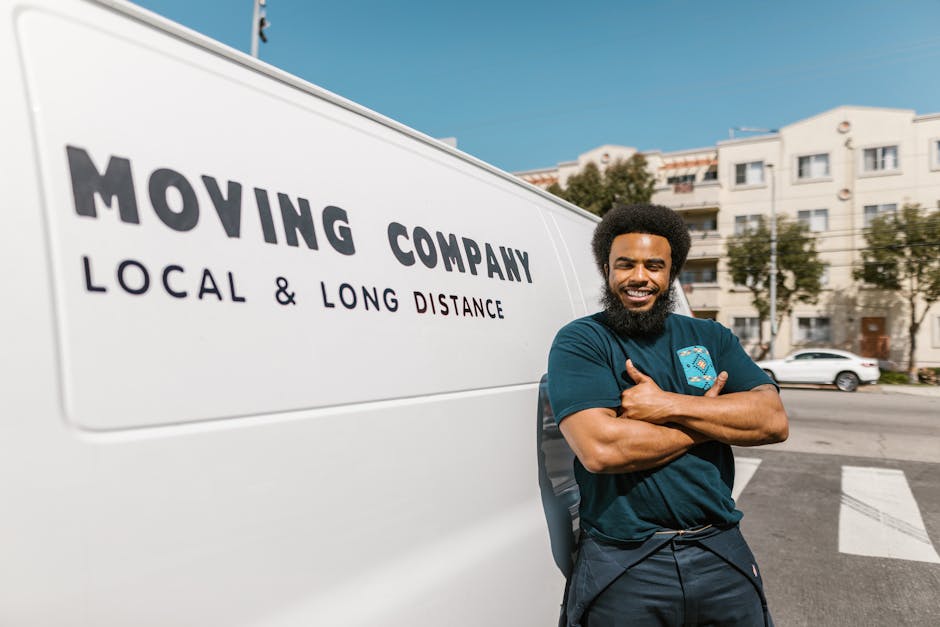 A smiling man with a curly afro hairstyle and beard, wearing a navy blue T-shirt with a moving company logo, stands outside next to a white moving van with the text 'MOVING COMPANY LOCAL & LONG DISTANCE' printed on its side. The man has his arms crossed and is positioned near the rear of the vehicle on a paved street with residential buildings and parked cars visible in the background. The scene is illuminated by natural daylight, highlighting the preparation for home relocation or furniture transport involving packing and loading processes as part of house removals, with the van ready for a move to or from the residence in Canonbury Lane, Highbury, as promoted by Highbury Man and Van.