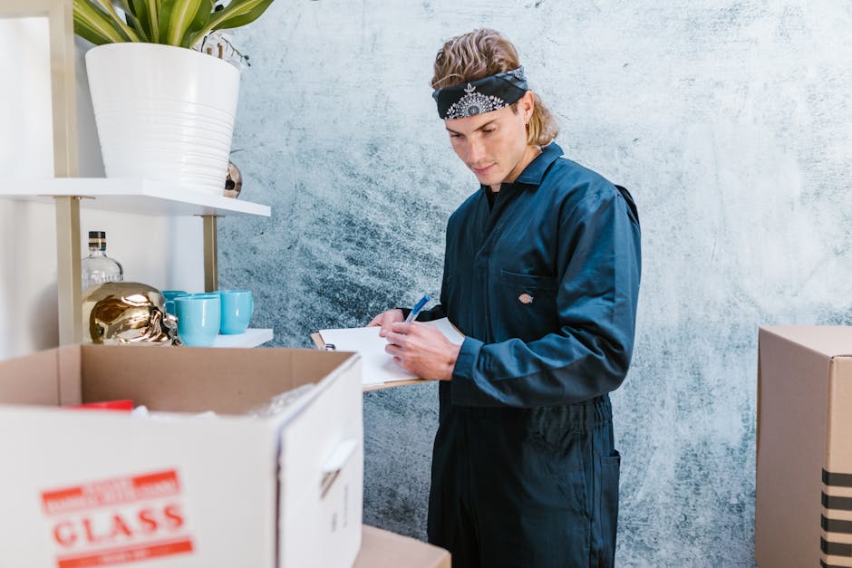 A young man wearing a dark blue work jumpsuit and a black bandana with white patterns is standing indoors next to a shelf with decorative items and storage jars. He is holding a pen and clipboard, possibly taking inventory or noting details during a home relocation or packing process. In front of him, there are several cardboard boxes, some partially open, containing packing materials like bubble wrap and small household items. The room has a textured light blue wall, and natural light illuminates the scene, highlighting the organized setup consistent with furniture transport and packing activities carried out by Highbury Man and Van as part of their removals service for a house move or relocation.