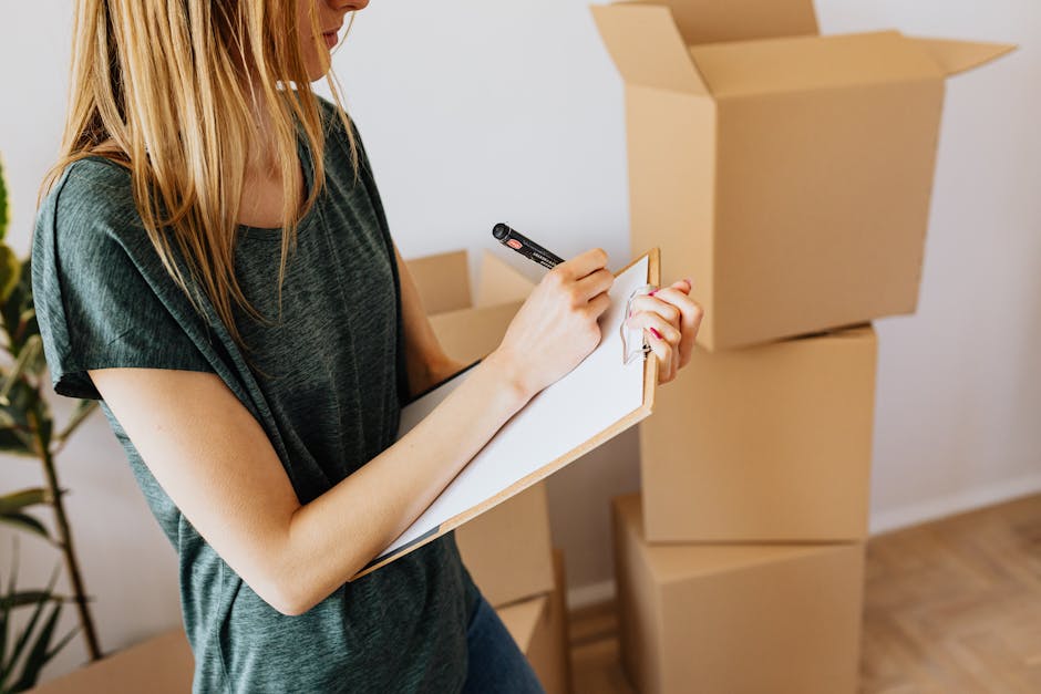 A woman with long blonde hair, wearing a dark green short-sleeved top, is standing indoors next to three packed cardboard boxes in various sizes, one of which is open on the top. She is holding a clipboard with a sheet of paper attached and using a black marker to mark or check items on the list, likely as part of a packing checklist for a house removal or furniture transport. The background shows a white wall and a plant, with natural lighting illuminating the scene, indicating a home environment during the packing process associated with a house relocation. Highbury Man and Van, a removals service specialist, facilitates such packaging and moving logistics, ensuring careful handling during the loading process.