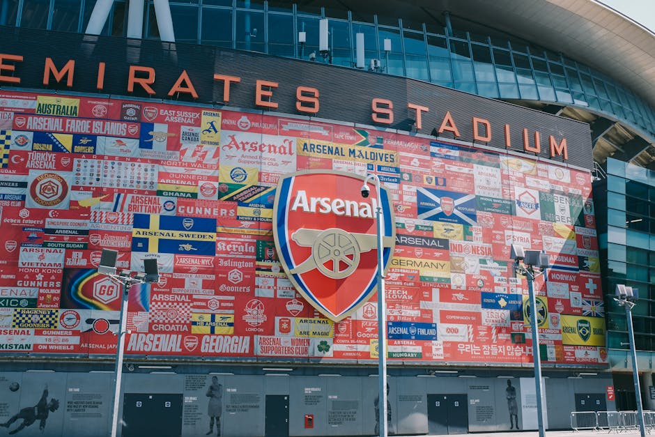 A large exterior wall of the Emirates Stadium featuring a prominent Arsenal Football Club emblem, surrounded by numerous smaller flags and banners representing different countries and regions, includi